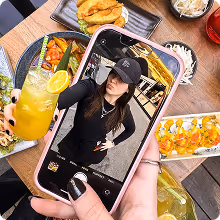 Phone displaying a woman in black outfit taking a selfie with food plates in the background